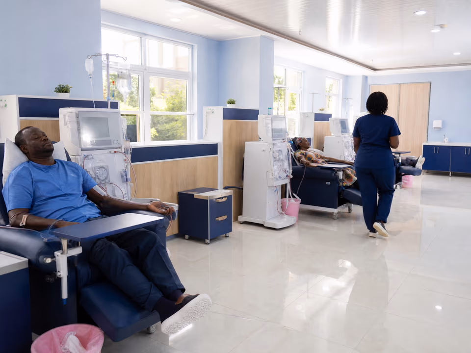 Illustrative scene: A bright dialysis unit with a row of machines and navy reclining chairs; a man rests in one while a nurse walks past.