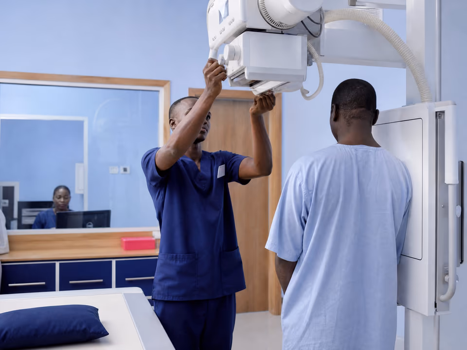 Illustrative scene: A radiographer in navy scrubs adjusts the X-ray tube for a man standing at the chest detector, seen from behind.