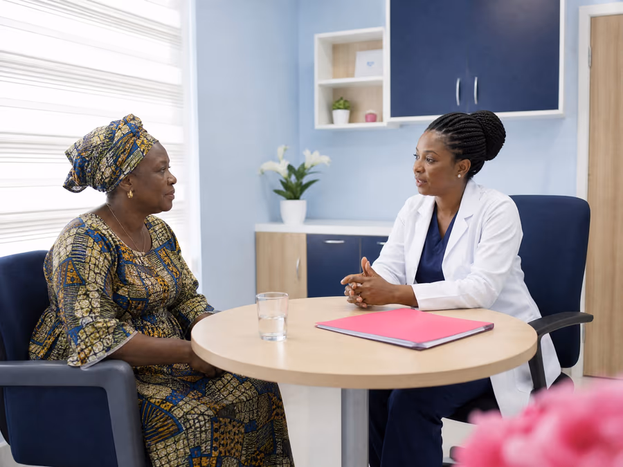 Illustrative scene: A woman in a headwrap talks with a clinician in a white coat at a round table in a bright consultation room.